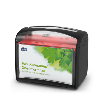 Stolní dávkovač ubrousků Tork Xpressnap® 272611 od společnosti TORK – Essity Professional Hygiene Germany GmbH má černou horní část a průhlednou přední část s designem zelených listů a nápisem „Tork Xpressnap One-at-a-time – lepší pro životní prostředí“. Tento dávkovač zlepšuje hygienu a zároveň efektivně snižuje spotřebu ubrousků.