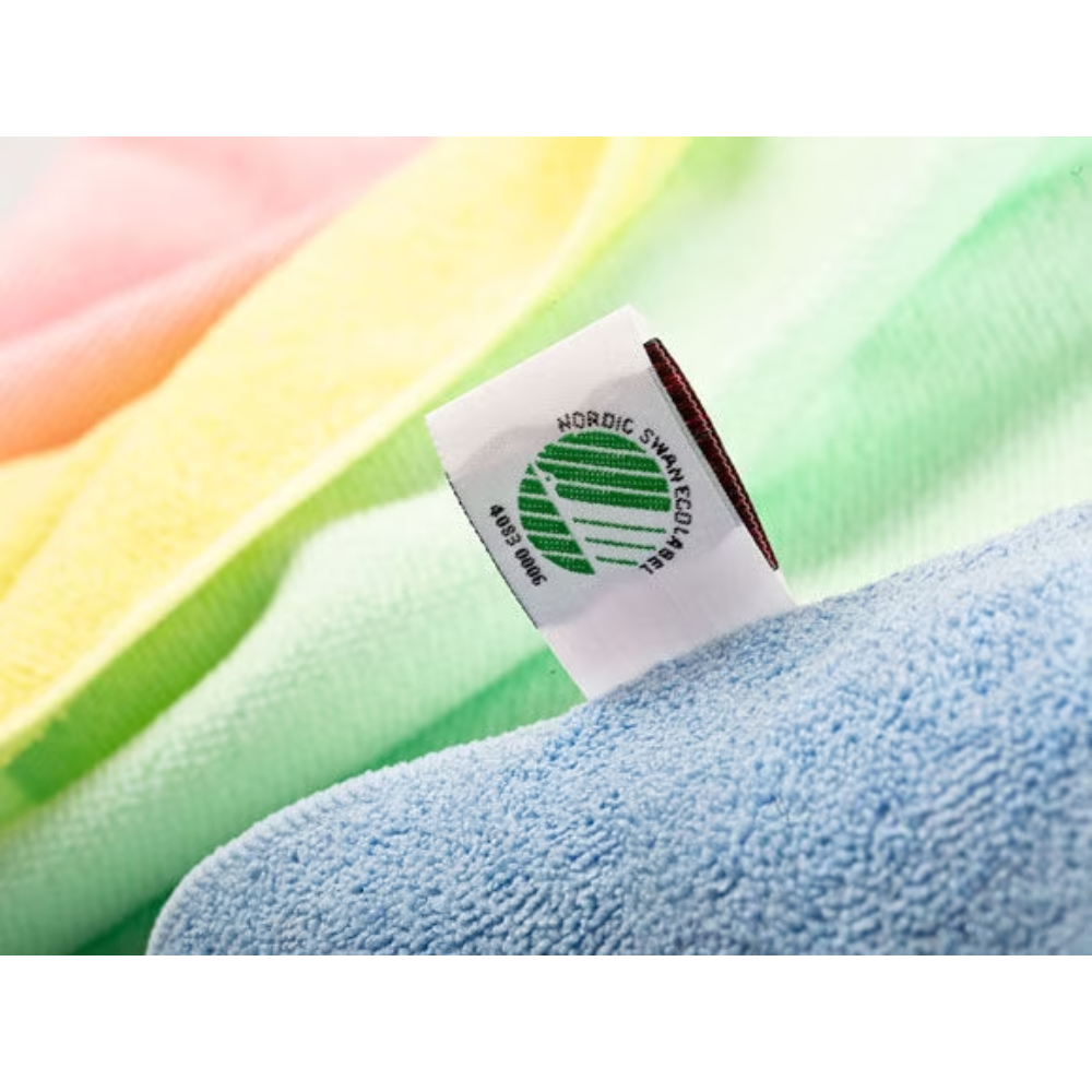 Detailní záběr na látkovou etiketu na ručníku Vileda Professional MicroTuff Plus se symbolem Nordic Swan Ecolabel. Ručník má jemné barevné tóny ve žluté, růžové, zelené a modré barvě s viditelnou texturou a je tedy ideální pro profesionální čištění. Je vyráběn společností Vileda GmbH a je známý jako udržitelná mikrovláknová utěrka.