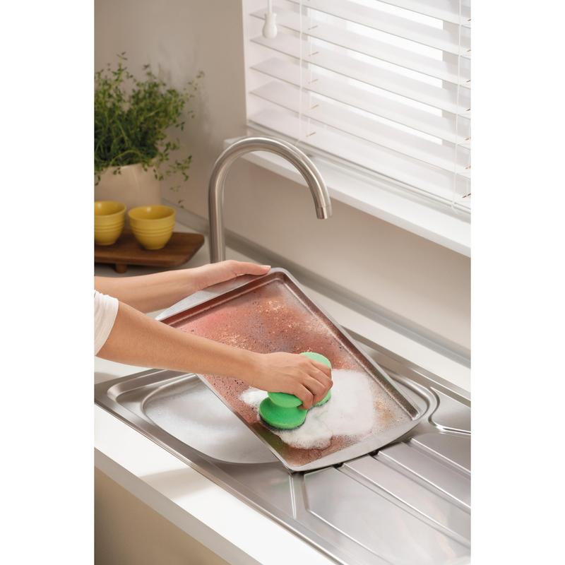 Někdo drhne velký, znečištěný plech s čisticí houbičkou Scotch-Brite® Classic od 3M Deutschland GmbH pod kohoutkem v světlé kuchyni s bílými žaluziemi, rostlinou a naskládanými žlutými miskami v pozadí.