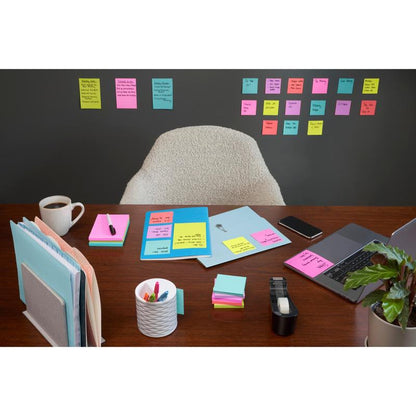 Uklizený stůl s laptopem, kávou, tužkami a kancelářskými potřebami stojí před zdí, která je pokryta řadami 3M Deutschland GmbH Post-it® Super Sticky Notes (47,6mm x 47,6mm, 90 listů/blok, 100% PEFC); za stolem stojí prázdná židle.