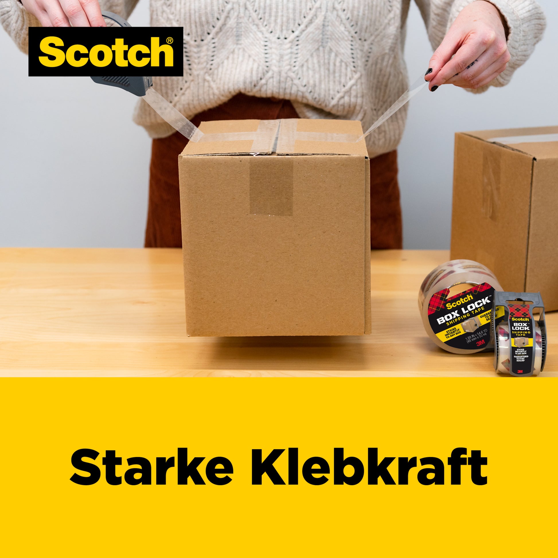 Eine Person verschließt eine Schachtel mit Scotch® Box Lock™ Verpackungsklebeband (48 mm x 20,3 m) von 3M Deutschland GmbH mit einem Spender. In der Nähe stehen ein weiterer Karton und weitere Rollen. Der Text lautet "Starke Klebkraft" neben dem Scotch-Logo.