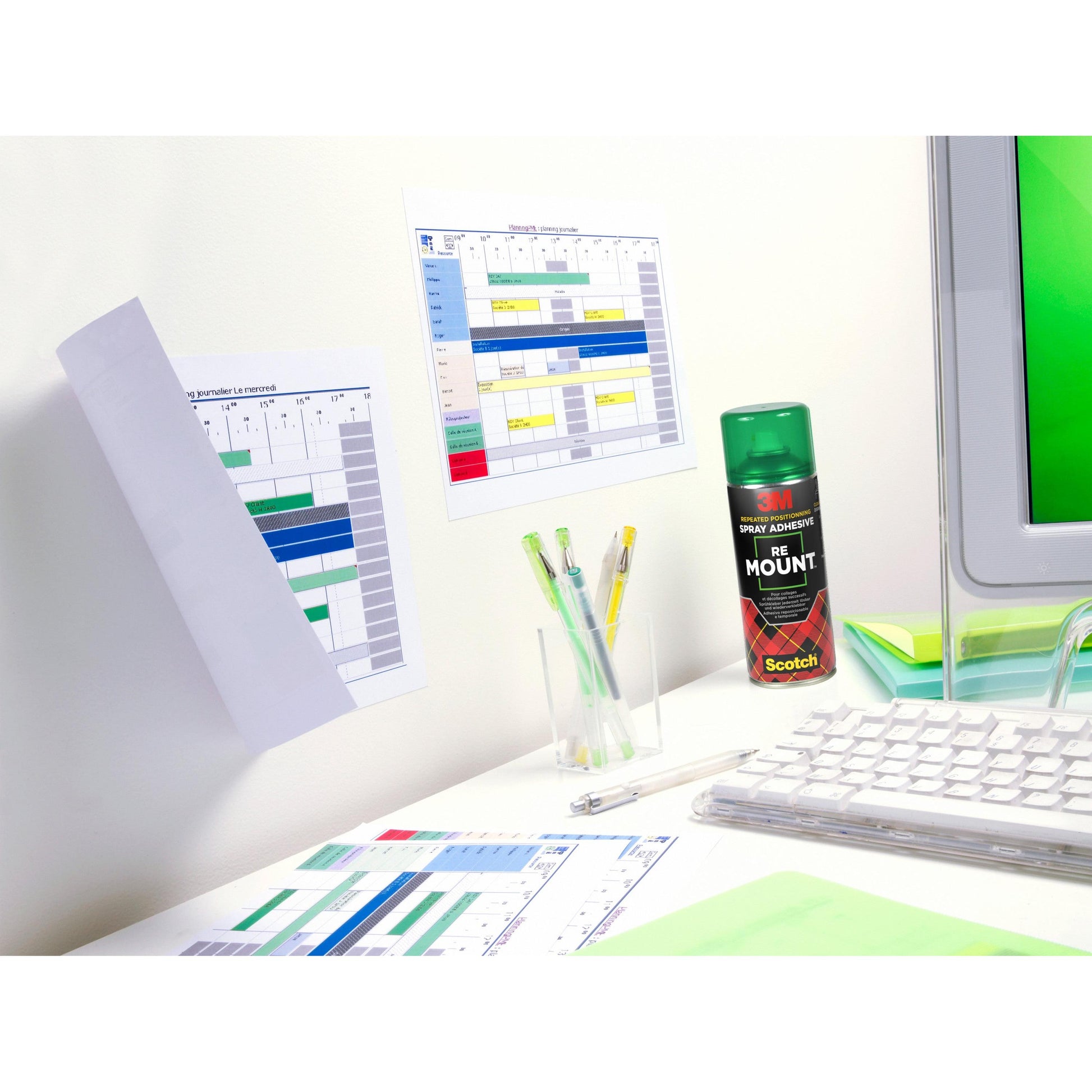 Ein Schreibtisch mit einer Tastatur, Stifte in einer durchsichtigen Halterung, gedruckte Zeitpläne an der Wand und auf dem Schreibtisch, ein Teil eines Monitors und eine Dose 3M™ Scotch® ReMount™ Klebespray (400 ml) der 3M Deutschland GmbH.