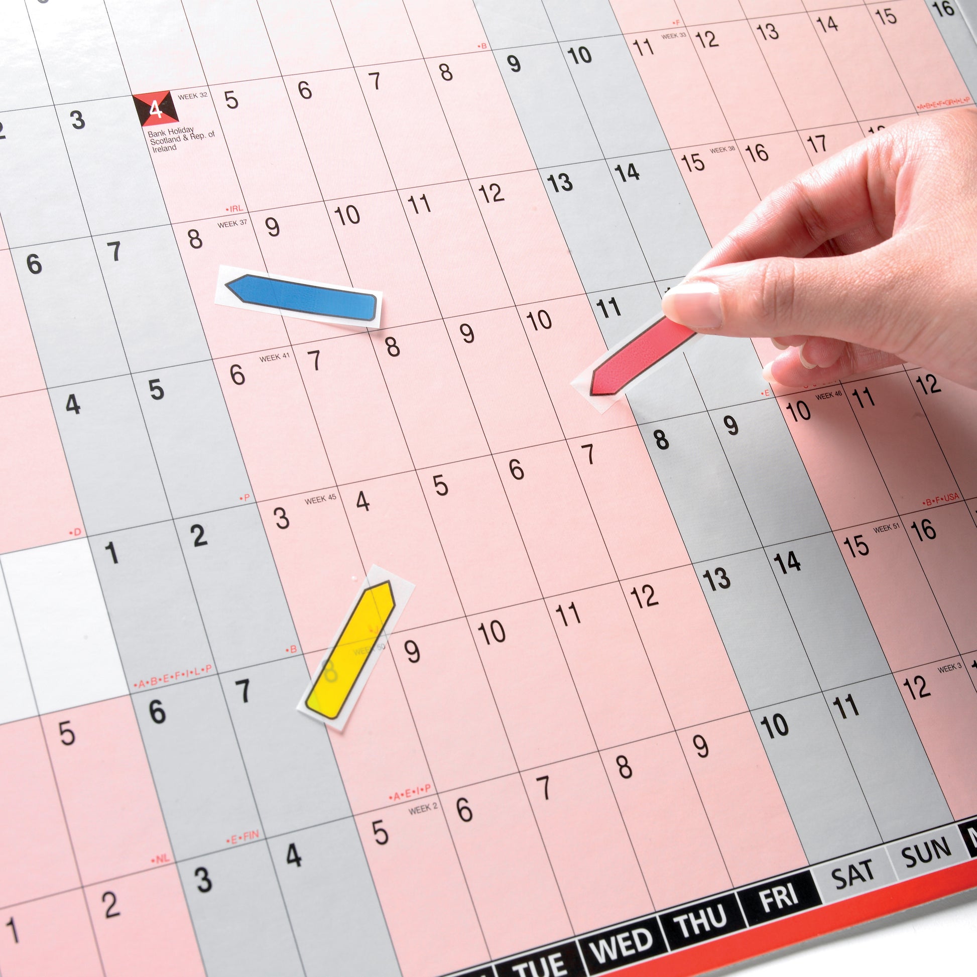Eine Hand platziert einen roten Post-it® Index Pfeil der 3M Deutschland GmbH auf einem großen Monatskalender, der auch gelbe und blaue Post-it® Pfeile enthält. Der Kalender zeigt Daten und Tage in grauen, rosa und weißen Quadraten an.